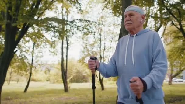 Старший дедушка в отставке практикует нордическую ходьбу на лыжных палках в летнем парке — стоковое видео