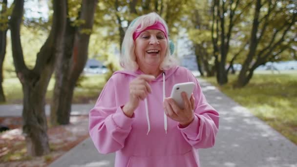 晨间公园，一名身穿耳机的田径高级女运动员正在收听智能手机的音乐 — 图库视频影像