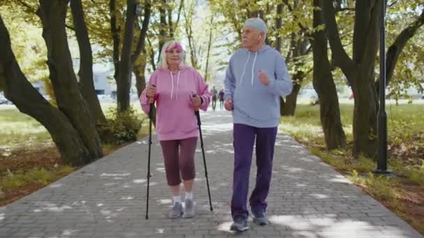 Активные бабушки и дедушки старших классов тренируют скандинавскую ходьбу с лыжными палками, бег в летнем парке — стоковое видео