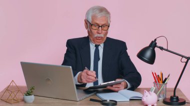 Oh my God Wow. Senior business man, using laptop, analysing paperwork documents raising hands in surprise, shocked by sudden victory, good news. Elderly grandfather freelancer working at pink office