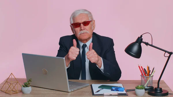 Senior funny business man in sunglasses using laptop computer notebook, raises thumbs up agrees with something or gives positive reply recommends advertisement likes good sits at desk at pink office