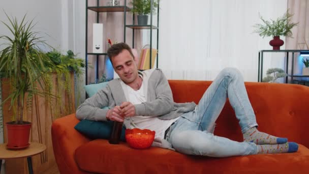 男人躺在沙发上的画像 吃薯片 喝啤酒 看有趣的电视连续剧 足球锦标赛 在家里在线社交媒体内容 享受家庭娱乐的男人 — 图库视频影像