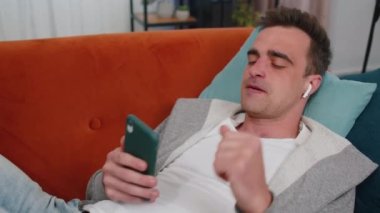 Kablosuz kulaklıklı mutlu beyaz adam evde koltukta dans ederek akıllı telefon uygulamasında en sevilen enerjik disko müziğini seçiyor ve içeride eğleniyor. İnsanlar hafta sonu aktiviteleri