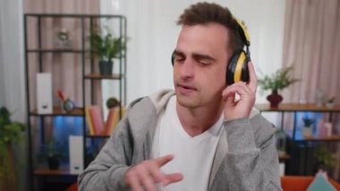 Kablosuz kulaklıklarıyla dans eden mutlu, rahat genç adam, oturma odasında şarkı söyleyen, mobil uygulamalarda favori enerjik disko müziğini seçen, kapalı alanda eğlenen. İnsanlar rahatlama aktiviteleri