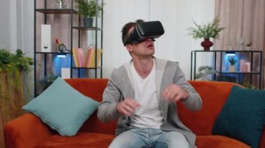 Şaşırmış bir adam evde VR kulaklık kullanıyor, 360 boyutlu video oyunu konseptinin tadını çıkarıyor, eğlendiriyor, olumlu bir ruh halinde, ellerini havada hareket ettiriyor, oturma odasında oturuyor. Sanal gerçeklik, modern aygıt