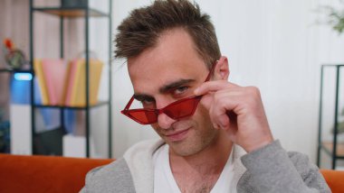 Kırmızı gözlüklü, baştan çıkarıcı, neşeli, stil sahibi, başarılı bir adamın portresi. Hoş bir gülümseme, göz kırpma, flört. Oturma odasında kanepede oturan genç, sevimli yetişkin bir adam.