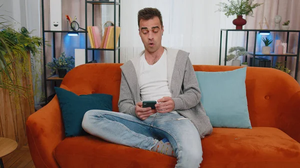 Oh my God Wow. Excited happy joyful winner man use smartphone typing browsing found out great big win good news celebrate victory. Young guy with mobile phone sitting at home in living room on couch