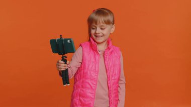Girl child toddler blogger taking selfie on mobile phone selfie stick, communicating video call online with subscribers. Young little children kid teen isolated alone on orange studio wall background
