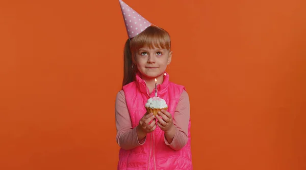 Happy one toddler children girl wearing festive cone cap celebrating birthday anniversary party blowing candle on small cake cupcake making a wish. Young little child kid isolated on orange background