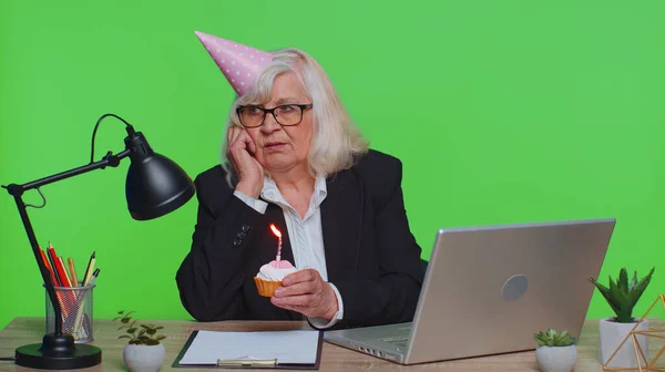Senior mature businesswoman celebrating lonely birthday in office, blowing candle on small cake making a wish. Elderly success grandmother woman wearing festive cap celebrate anniversary party alone