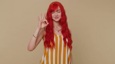 Neşeli, mutlu, kızıl saçlı kadın kameraya onaylı geri bildirim jesti göstererek bakıyor. Bej renkli stüdyo geçmişi olan kızıl saçlı genç bir kız. İnsanlar samimi duygular