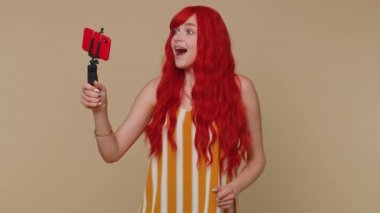Vay canına. Etkilenmiş kızıl saçlı kadın blogcu cep telefonu özçekim çubuğuyla selfie çekiyor, abonelerle online video görüşmesi yapıyor. Kızıl saçlı genç kız bej renkli stüdyo arka planında tek başına.