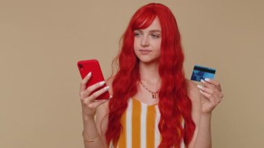 Samimi kızıl saçlı kadın müşteri kredi kartı ve akıllı telefon kullanarak para aktarıyor, internetten alışveriş yapıyor, ödeme yapıyor. Finans, internet. Genç kız bej duvar arkasında tek başına.