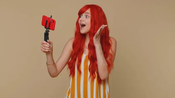 Wow reaction. Impressed redhead woman blogger taking selfie on mobile phone selfie stick, communicating video call online with subscribers. Young girl ginger isolated alone on beige studio background