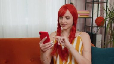 Kanepede oturan neşeli kadın modern ev dairesinde cep telefonu tebessümü kullanıyor. Mutlu kızıl kız akıllı telefon sosyal medya uygulamaları üzerinden mesajları paylaşıyor, rahatlama filmi izliyor