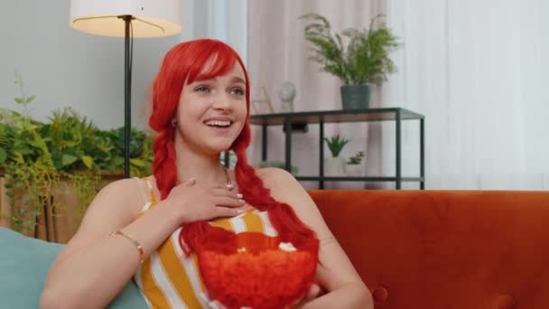 兴奋的红头发年轻女子坐在沙发上吃爆米花 看有趣的电视连续剧 体育锦标赛游戏 在线社交媒体内容 生姜女孩享受家庭娱乐 — 图库视频影像
