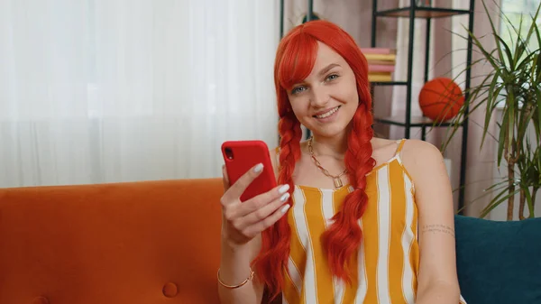 坐在沙发上快乐的女人在现代化的家庭公寓里用手机微笑 快乐的红头发女孩在智能手机社交媒体应用程序上发短信 分享内容 观看轻松电影 — 图库照片