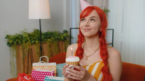 Lovely redhead woman wears festive birthday hat hold cupcake makes wish joyful congratulating blowing burning candle on cake. Young ginger girl celebrating anniversary party on sofa with gifts at home