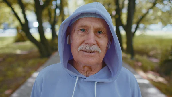 Portrait of athletic fitness senior elderly sport runner man training, listening music in earphones, wearing hood, looking at camera and smiling. Grandfather workout cardio outside in park at morning