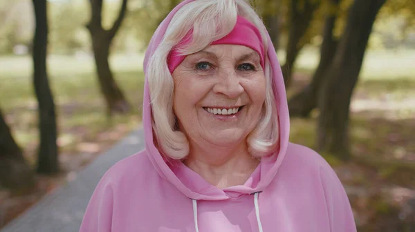 Portrait of athletic fitness senior elderly sport runner woman training, listening music in earphones, wearing hood, looking at camera, smiling. Grandmother workout cardio outside in park at morning