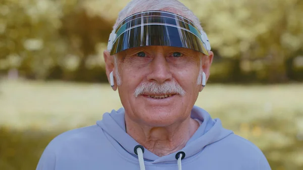 Portrait of athletic fitness senior elderly sport runner man training, wearing earphones listening music, looking at camera and smiling. Healthy grandfather workout cardio outdoors in park at morning
