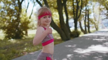 Atletik fitness sporunun arka görünüşü, yürümeye başlayan koşucu kızın maraton antrenmanına, yarışa başparmak göstererek başlamasına neden oldu. Parkta kardiyo çalışması. Çocuk koşunun tadını çıkarıyor. Aktif çocuk koşucusu