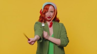 Yemek hazır. Yeşil ceketli ve elbiseli, çatal bıçaklı yemek servisini bekleyen aç, kızıl saçlı genç kadın lezzetli restoran yemeklerini takdir edecektir. Kızıl kız sarı arka planda izole edilmiş.