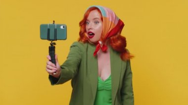 Vay canına. Etkilenmiş kızıl saçlı kadın blogcu cep telefonu özçekim çubuğuyla selfie çekiyor, abonelerle online video görüşmesi yapıyor. Kızıl saçlı genç kız sarı stüdyo arka planında tek başına izole edilmiş.