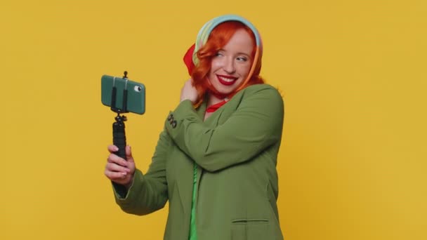 ジャケット旅行ブロガーの赤毛の女性は 携帯電話の自撮り棒に自撮りを取り 加入者とオンラインでビデオ通話を通信します 若いです生姜女の子孤立した一人で黄色のスタジオ背景 — ストック動画