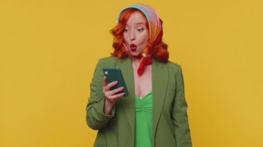 Akıllı telefon görünümlü mutlu kızıl saçlı genç kadın içten bir sevinç gösterisi kazanıyor, para dolar banknotları alıyor, başarı piyangosunda şans getiriyor. Stüdyodaki kızıl kız sarı arka planda izole edilmiş.