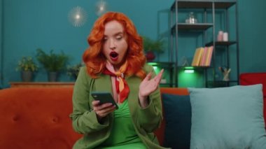 Aman Tanrım. Vay canına. Heyecanlı ve neşeli kazanan kadın akıllı telefon daktilo kullanıyor. Kazanan kazanır. İyi haber zaferi kutlar. Cep telefonlu genç kızıl kız evinde, kanepede oturuyor.