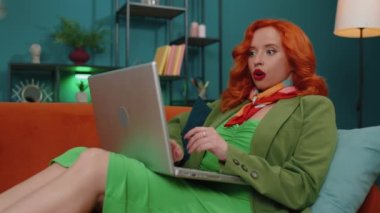 Aman Tanrım. Vay canına. Şaşırmış kızıl saçlı genç kadın dizüstü bilgisayar kullanıyor, iyi haber mesajı alıyor, ani zaferi kutluyor, galibiyeti kutluyor, evden online alışveriş yapıyor. Oturma odasındaki kanepede oturan kızıl kız.