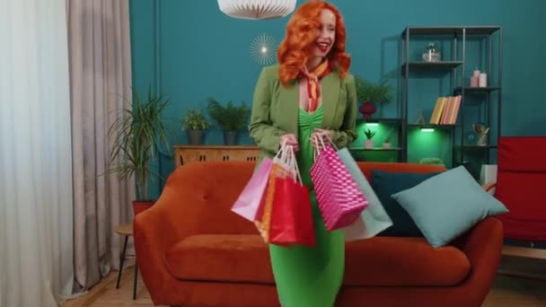 緑の服を着た幸せな赤毛の若い女性のお店の消費者は 現代のアパートで袋で買い物をした後 家に戻ってきました 満足した生姜の女の子は リビングルームでオンライン注文から小包を受け取りました — ストック動画