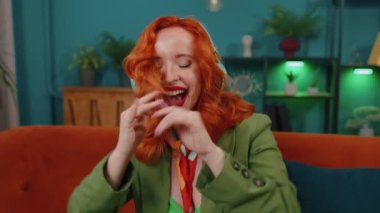 Kablosuz kulaklıklı mutlu, rahat, kızıl saçlı kadın dans ediyor, oturma odasında şarkı söylüyor, mobil uygulamalarda favori enerjik disko müziğini seçiyor, kapalı mekanlarda eğleniyor. Kız hafta sonu aktiviteleri