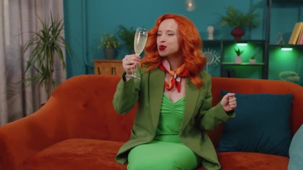喜びに満ちた赤毛の女性は シャンパンの応援と飲酒のガラスを保持し 成功を祝い キャリアが成長し リビングルームで自宅で幸運な状況を祝います 新しい家の家賃や購入を祝うジンジャーガール — ストック動画