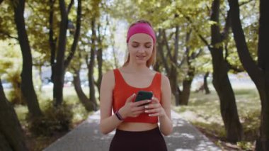 Sporcu spor koşucusu kız yürüyor ve kablosuz kulaklıklarla akıllı telefon dinliyor. Şehir parkında egzersiz kardiyosu. Genç kadın koşucu sağlıklı yaşam tarzını seviyor.