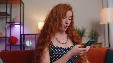 Aman Tanrım. Vay canına. Heyecanlı ve neşeli kazanan kadın akıllı telefon daktilo kullanıyor. Kazanan kazanır. İyi haber zaferi kutlar. Cep telefonlu genç kızıl kız evinde, kanepede oturuyor.