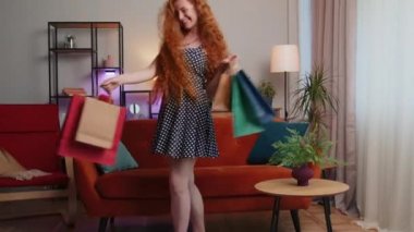 Polka puantiyeli elbise giyen mutlu kızıl saçlı tüketici, modern dairede çanta sattıktan sonra eve geri döndü. Tatmin olmuş kızıl kız oturma odasında internetten paket aldı.