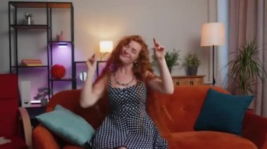 Moda, neşeli, pozitif saçlı, dans eden, ritme ayak uyduran, el kaldıran, modern ev dairesinde dublaj yapan genç bir kadın. Oturma odasındaki kanepede oturan kızıl güzel kız.