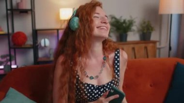 Kablosuz kulaklıklı mutlu, rahat, kızıl saçlı kadın dans ediyor, oturma odasında şarkı söylüyor, mobil uygulamalarda favori enerjik disko müziğini seçiyor, kapalı mekanlarda eğleniyor. Kız hafta sonu aktiviteleri