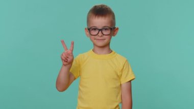 Mutlu gülümseyen küçük çocuk zafer işareti gösteriyor, başarı umuduyla barış jesti yapıyor, iyimser bir gülümsemeyle gülümsüyor. Genç anaokulu çocuğu mavi stüdyo geçmişinde izole edilmiş.