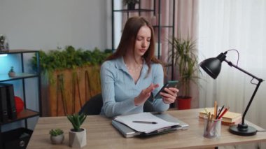 Serbest çalışan genç bir kadın akıllı telefon sosyal medya uygulamaları üzerinden mesajlarını paylaşıyor. İnternetten alışveriş alışverişi yapıyor. Ev ofisinin masasında oturuyor. Kız öğrenci çalışma alanı iş eğitimi