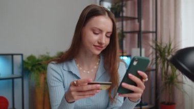 Kredi kartı ve akıllı telefon kullanan genç bir kadın para transfer ederken internetten alışveriş yapıyor. Ev ofisinin masasında oturuyor. Uzaktan kumanda işinde çalışan kız öğrenci.
