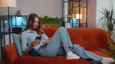 Genç bir kadın para transfer ederken kredi kartı ve akıllı telefon kullanıyor, online alışveriş yapıyor, modern bir evde yemek siparişi veriyor. Oturma odasında kanepede uzanan güzel kız.
