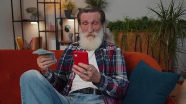 Aile bütçesi planlamak. Gülümseyen yaşlı büyükbaba nakit para sayıyor, akıllı telefon kullanıyor ev faturalarını hesaplıyor. Neşeli adam gelirinden memnun ve planlı tatil ve hediyeler için para biriktiriyor.
