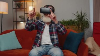 Yaşlı büyükbaba, sanal gerçeklik teknolojisi VR uygulama kulaklığı kullanarak video oyunu simülasyonu yapıyor ve modern ev dairesinde film izliyor. Gözlüklü adam kanepede oturuyor.