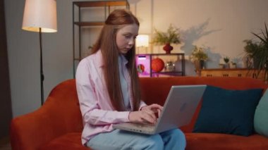 Young redhead child girl sits on sofa closing laptop pc after finishing work in living room at home. Red hair freelancer working online, remote job. E-learning, browsing internet on notebook computer
