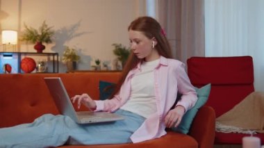 Young redhead teenager child girl freelancer at home room couch working on laptop. Ginger children kid works on notebook, sends messages, makes online order purchases or food delivery, watching movies