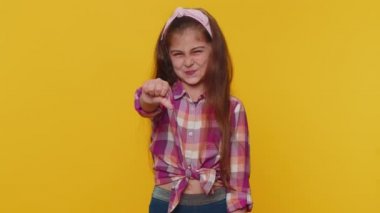 Dislike. Upset young preteen child girl kid showing thumbs down sign gesture, expressing discontent, disapproval, dissatisfied bad work indoors. Displeased toddler children on yellow studio background