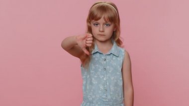 Dislike. Upset young preteen child girl kid showing thumbs down sign gesture, expressing discontent, disapproval, dissatisfied bad work indoors. Displeased toddler children on pink studio background